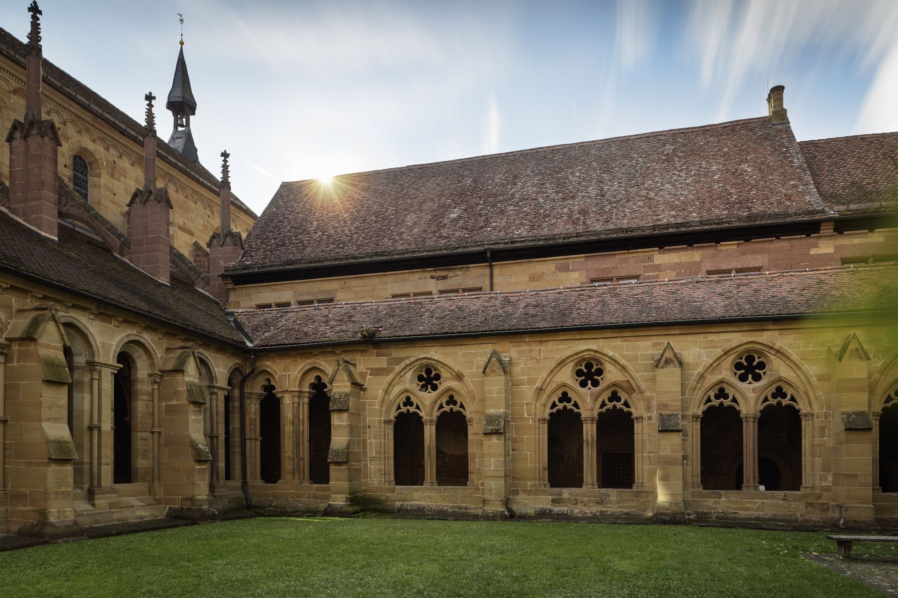 Gebäude aus Sandstein mit großen Maßwerkfenstern, dahinter die Klosterkirche